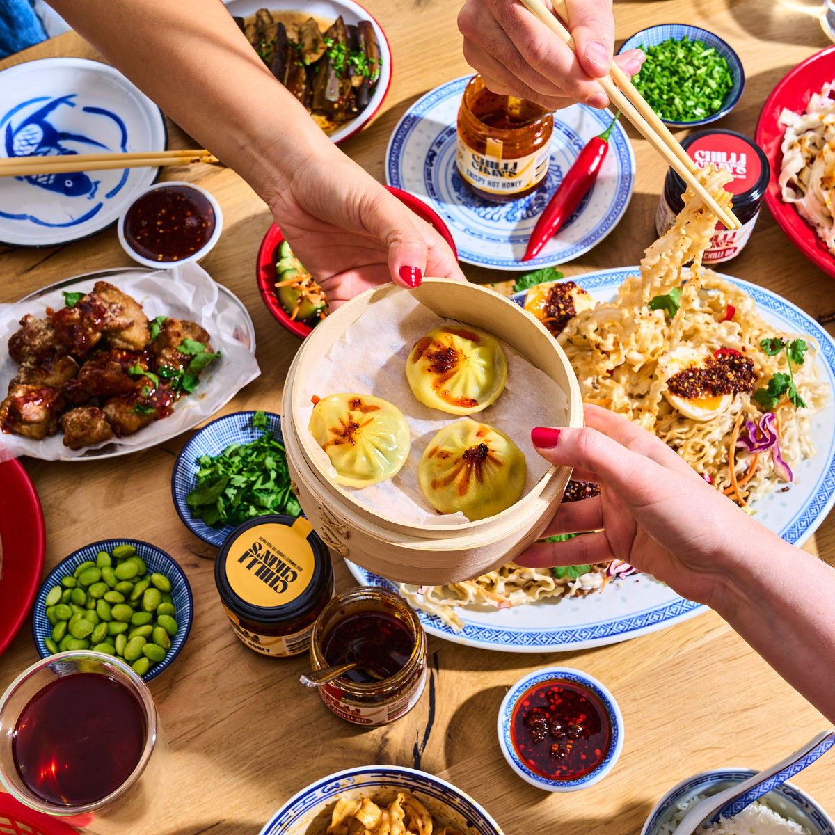 Hands sharing steamed dumplings and a bowl of knife-cut noodles with Chilli Chan's jars on the table